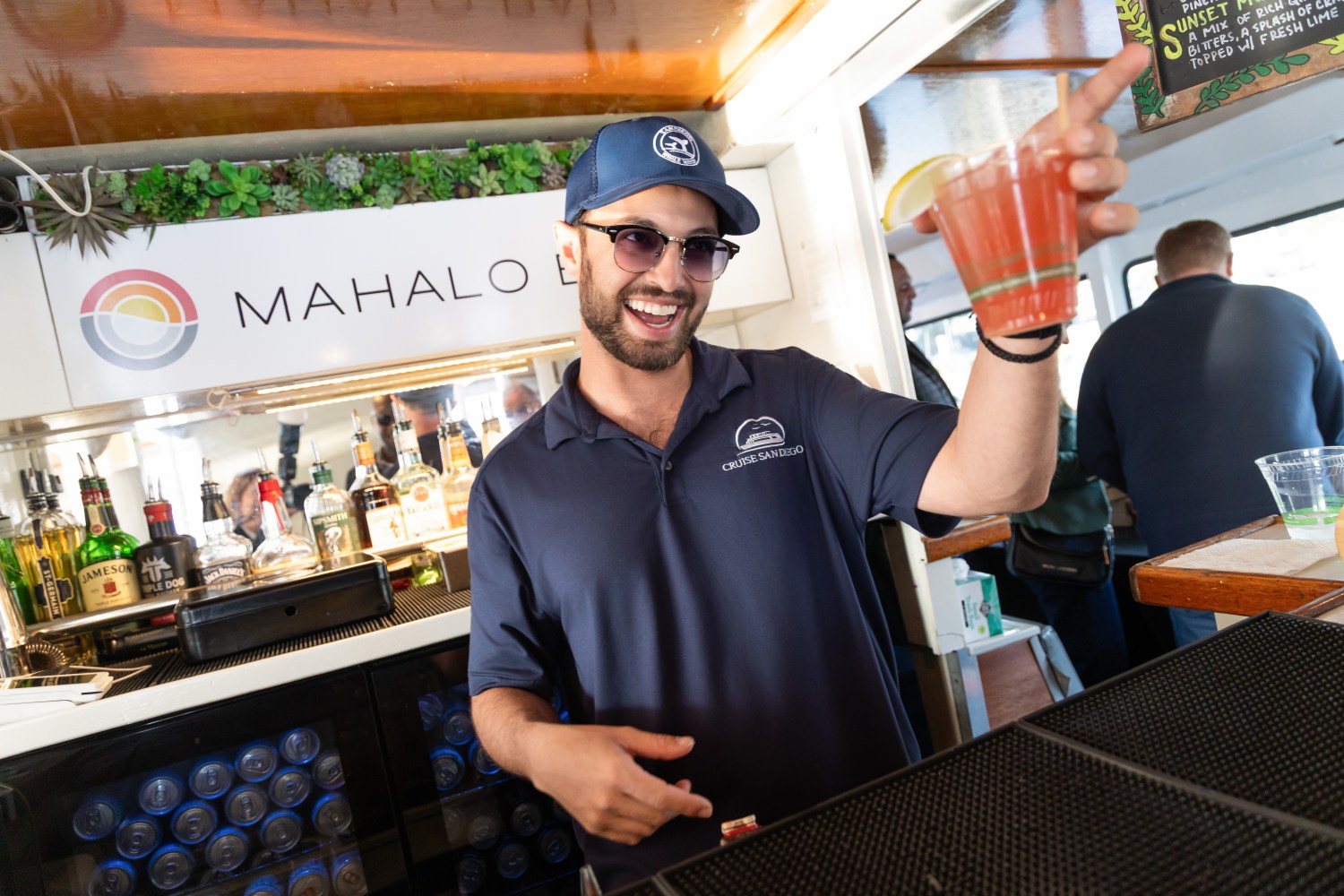 Smiling bartender holding fruity drink at boat bar, wearing sunglasses and cap.