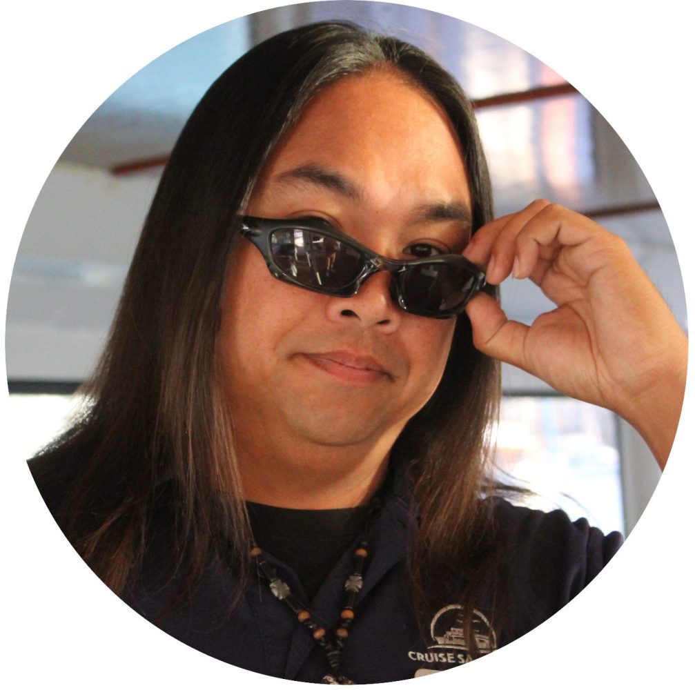 Person with long hair adjusting sunglasses, wearing a dark shirt and necklace.