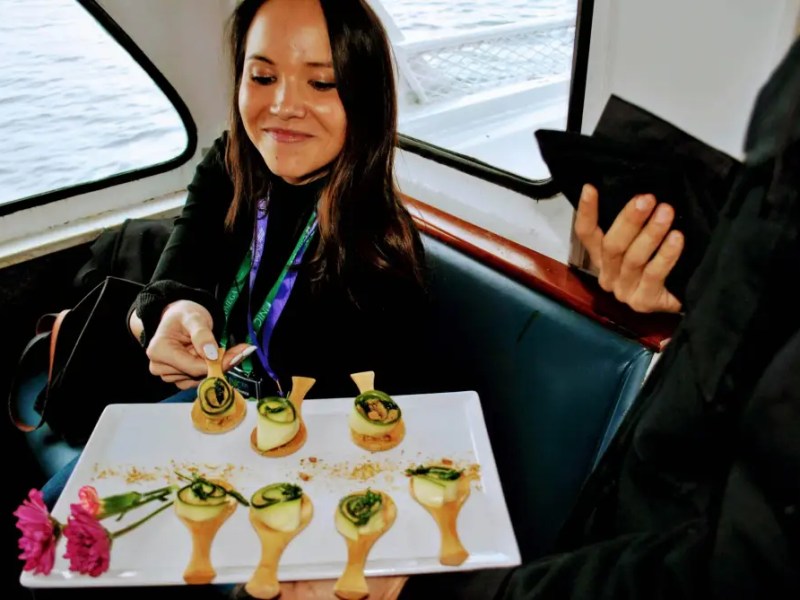 Woman on a boat holding a platter with appetizers and flower, smiling at another person.