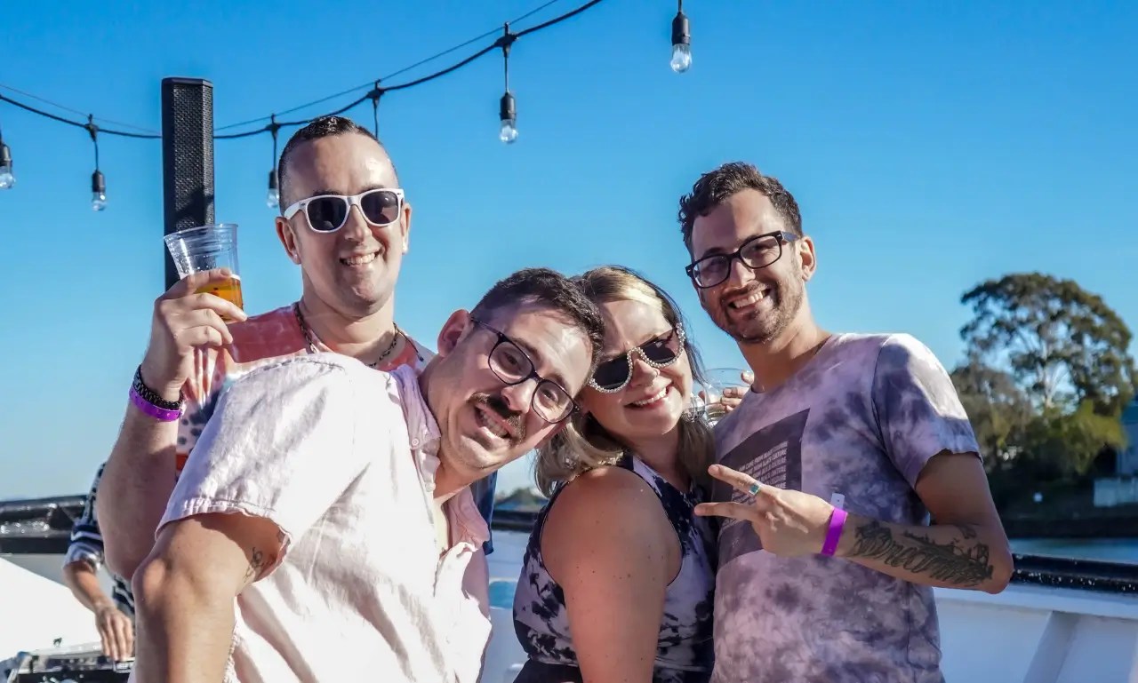 Four people smiling and posing outdoors under string lights on a sunny day.