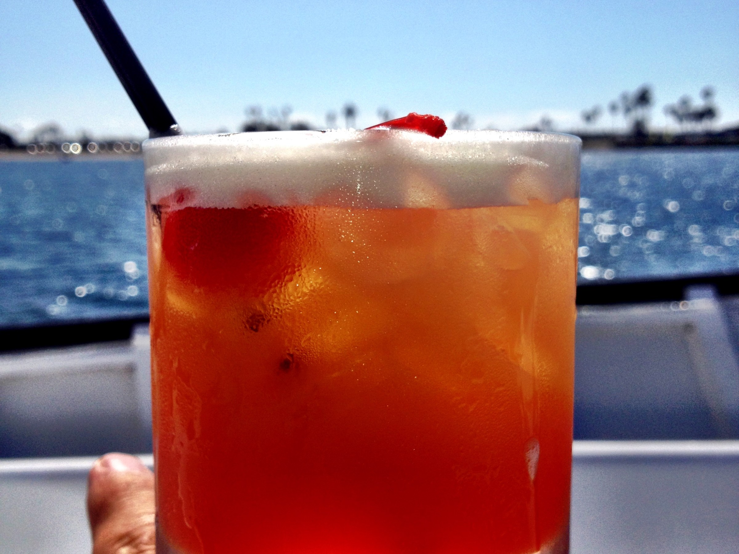 Hand holds a cocktail with a straw by the water on a sunny day.