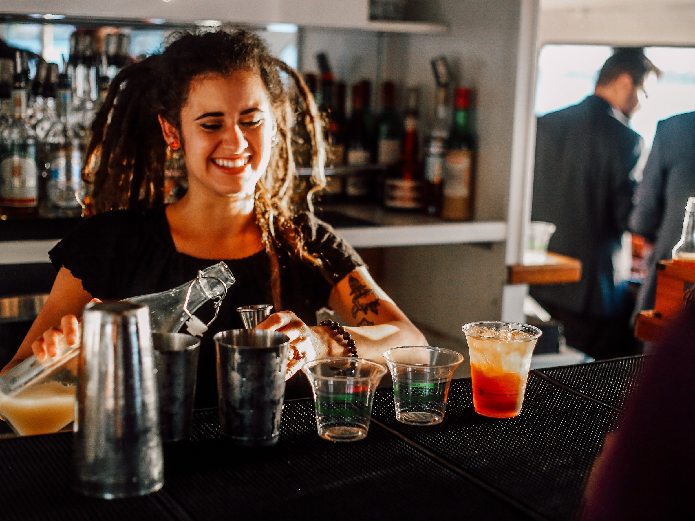 Bartender with dreadlocks pours drinks at a bar.