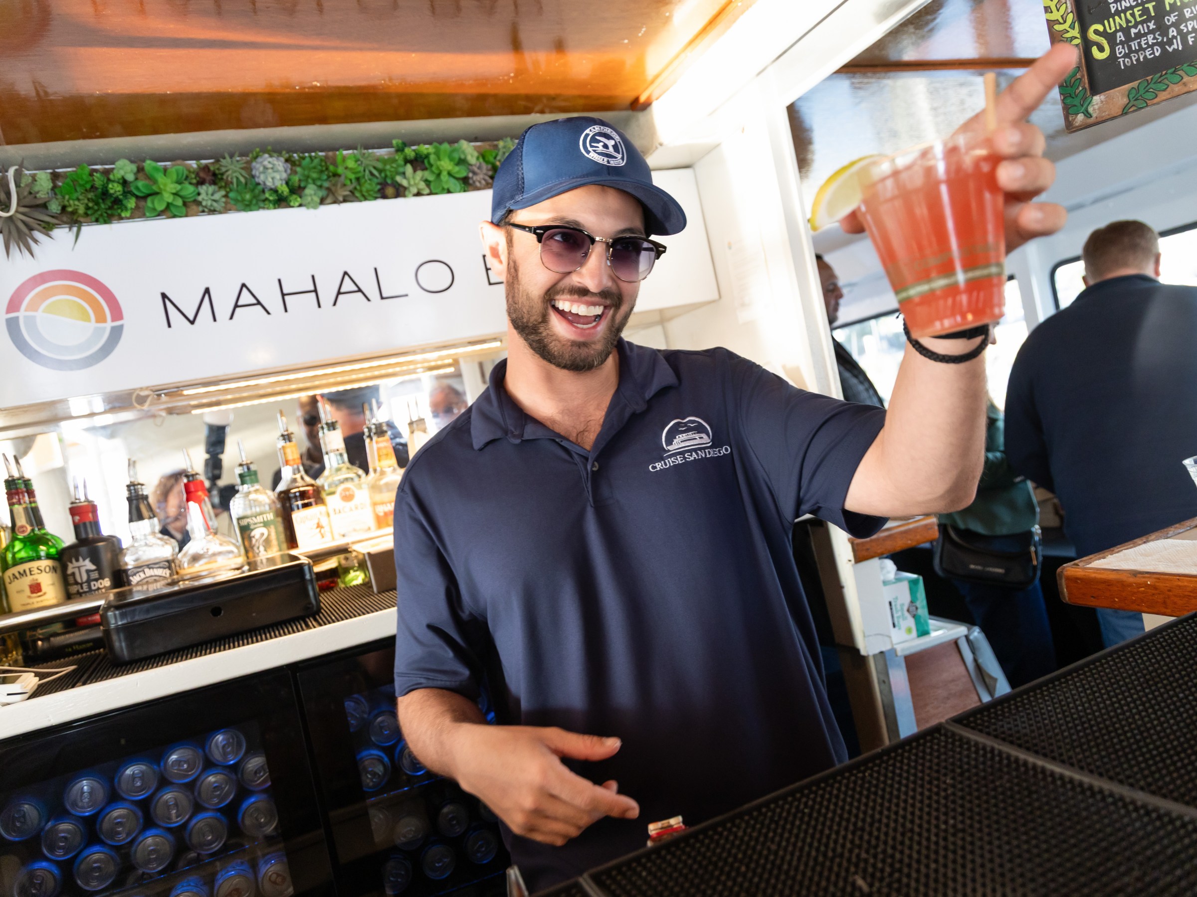 Smiling man behind bar holding red drink, wearing sunglasses and cap.