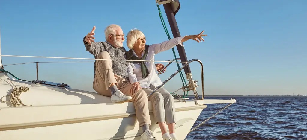 Elderly couple sitting on a sailboat, smiling and pointing at the sea.